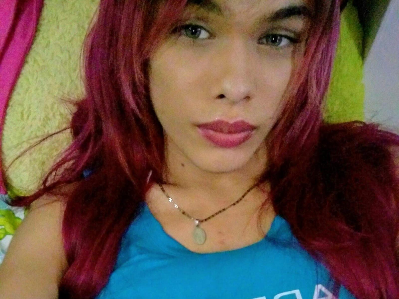 LaddyMorningstar, con i capelli rossi, una canottiera blu, rossetto rosa e un piccolo ciondolo, sdraiata, che guarda la fotocamera con le labbra leggermente arricciate..