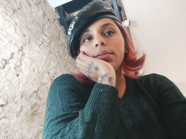 Chili's leaning against a stone wall, hand on cheek, wearing a black cap and green sweater, red hair, tattoos visible.