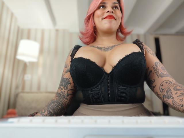 Chili with red bob, black lace corset, and tattoos, leaning forward, showing cleavage, red lipstick, and confident gaze.