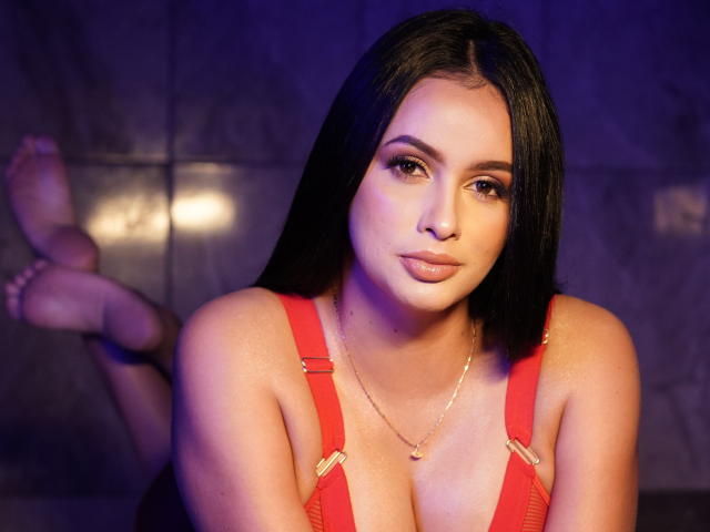 EmilyParkerr with long black hair, red suspenders, and a gold necklace, has her tits visible, legs up, and a sultry expression, in a purple-lit photo.