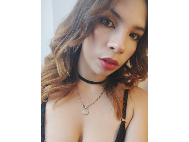 VioletaStone with wavy brown hair, black choker, and black lace bra, showing her tits, wearing a silver necklace, and red lipstick, staring at the camera.