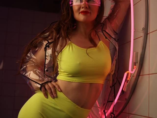 DashaHart with long brown hair, wearing a neon yellow sports bra and matching high-waisted pants, transparent plastic jacket, pink neon glasses, and red lipstick, leaning on a white tiled wall.