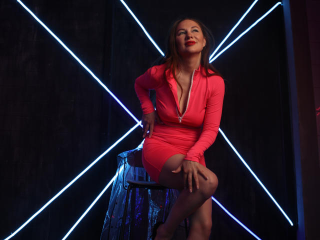 DashaHart in un abito rosa aderente con zip, seduta su uno sgabello, gambe incrociate, mani sulle ginocchia, linee blu neon sullo sfondo, rossetto rosso, sorriso sicuro, capelli castani..