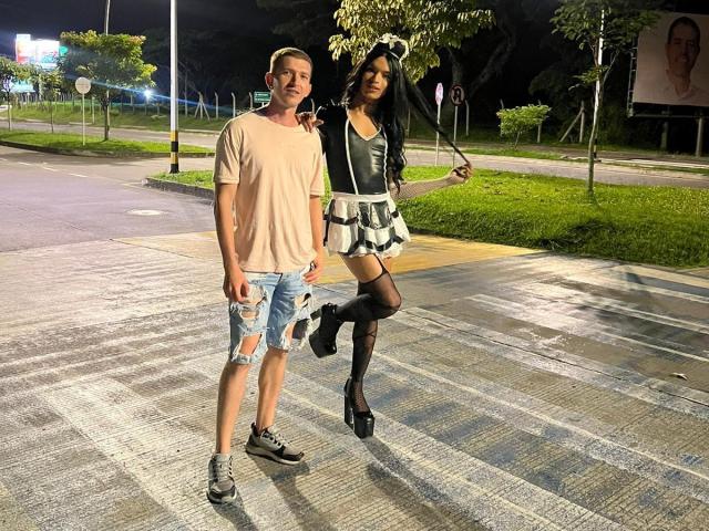 CoupleSensualHot with black hair, long legs, and black thigh-highs in a frilled skirt, stands next to a guy in a beige tee and ripped shorts, both on a wet sidewalk.