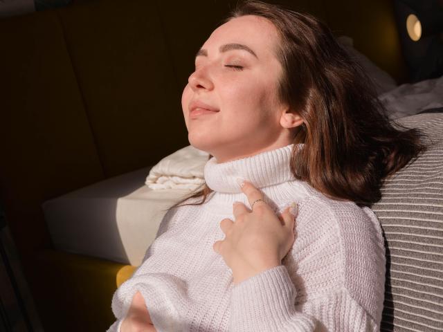 Aimeekai with closed eyes, wearing a white turtleneck, sunlight on her face, hand on chest, relaxed, brown hair, slight smile, soft skin, sitting on a striped chair.