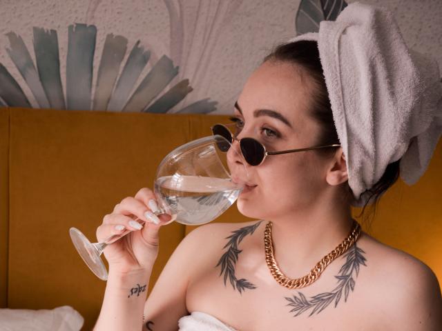 Aimeekai, avec une serviette sur la tête, portant des lunettes de soleil rondes, buvant dans un verre de vin, exhibant sa poitrine avec des tatouages de feuilles et un collier en chaîne en or..