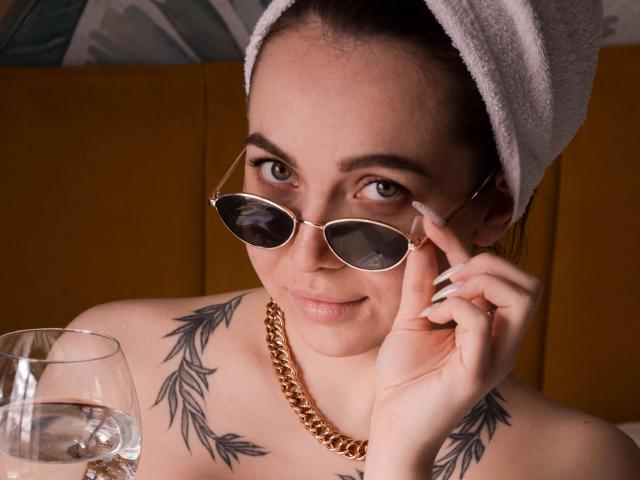 Aimeekai, avec une serviette sur la tête, portant des lunettes de soleil aviateur, une chaîne en or et des tatouages de feuilles sur la poitrine, tenant un verre d'eau, souriant. Ses seins sont visibles..
