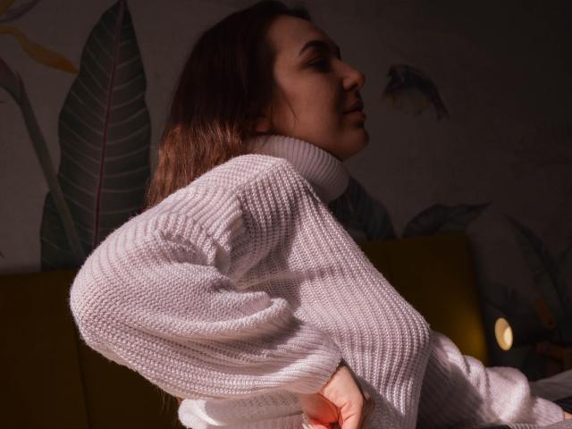Aimeekai with long brown hair, wearing a white knit turtleneck, side profile, hand on hip, soft lighting, subtle smile, green leaf pattern in the background, cozy atmosphere.