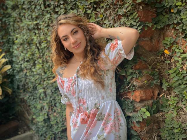 LuciaPerth with wavy blonde hair, wearing a floral dress, stands against a brick wall covered in green ivy, smiling with one hand in her hair, showing her cute tits.