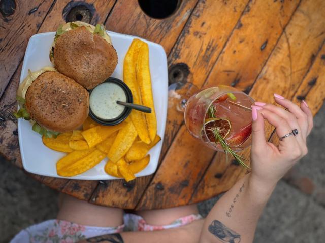 As mãos de LuciaPerth, com unhas rosadas e um anel, segurando uma bebida com ervas, acima de uma mesa de madeira com hambúrgueres, batatas fritas amarelas e um molho, em um prato branco..