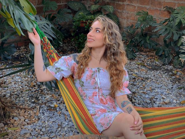 LuciaPerth, con i capelli biondi ricci, un vestito floreale, seduta in un'amaca colorata, sorridente, che tiene una foglia, con un tatuaggio visibile sul braccio destro, su un terreno di ghiaia, con piante verdi..