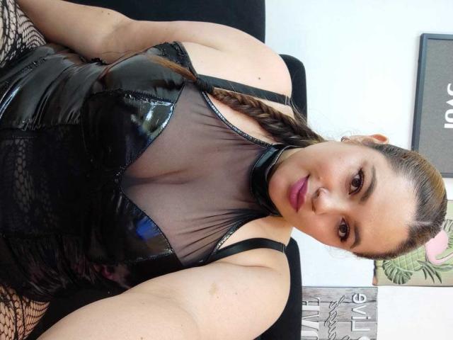 LissaClaire with braided brown hair, black glossy vinyl top, sheer mesh, and pink lipstick, showing her tits, sitting on a black chair, fishnet on lower left.