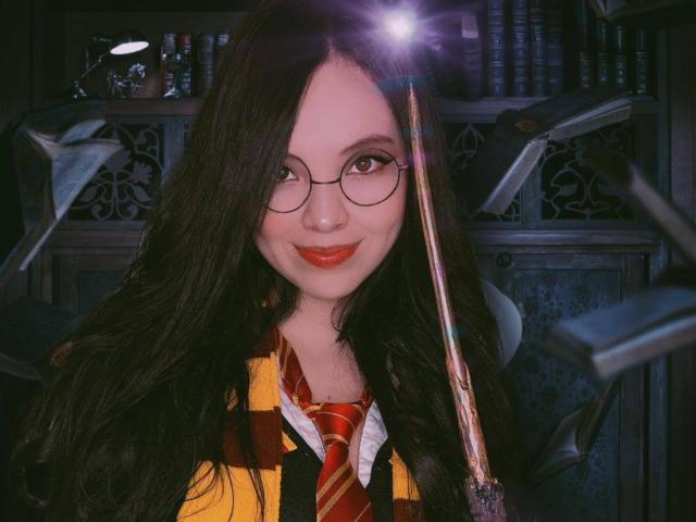 ElyMoon met lang zwart haar, ronde bril, rode lippenstift, een geruit shirt in geel en rood, die een toverstaf vasthoudt, lachend, met een boekenkast op de achtergrond, zacht licht, schattig, nerdy, sexy..