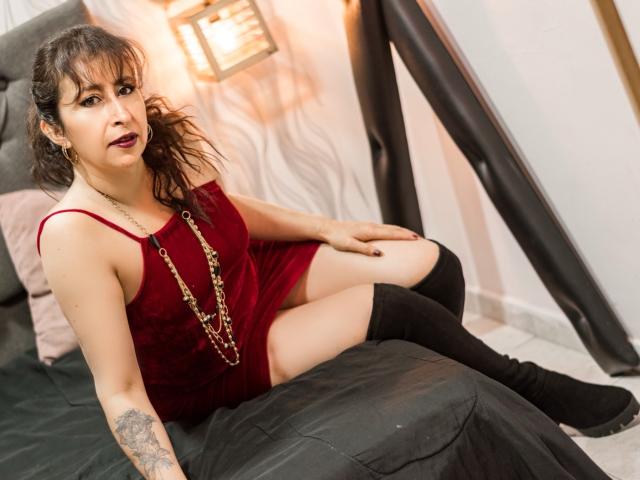 Viktoria Brown, mit dunklen, welligen Haaren, einem roten Samtkleid und schwarzen, hohen Stiefeln, sitzt auf einem schwarzen Bett, zeigt ihren tätowierten Arm und ihren kleinen Hintern und trägt eine Halskette, mit dunklem Lippenstift..