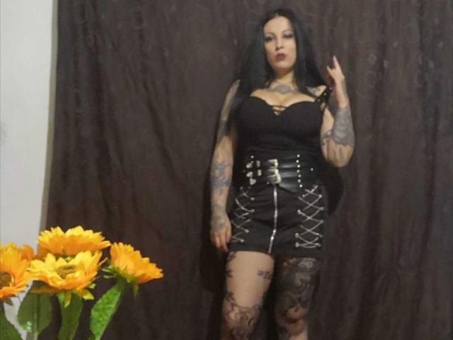 Astartex, con i capelli neri, tatuaggi e rossetto scuro, indossa un top nero senza spalline e una gonna con lacci, tiene un dito alzato, ed è in piedi accanto a girasoli..