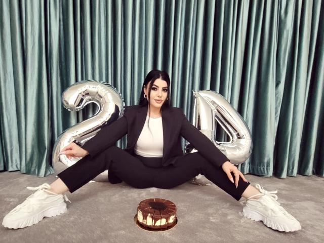 MelissaGlow sits with legs spread, wearing a black suit, white top, and white sneakers. She's between shiny "20" balloons, with a chocolate donut in front.