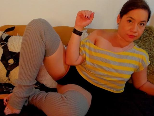 MsJustineBeaux, vestita con una camicia a righe gialle e grigie, pantaloncini neri e scaldamuscoli grigi, è seduta su un divano con una gamba sollevata, mostrando il sedere..
