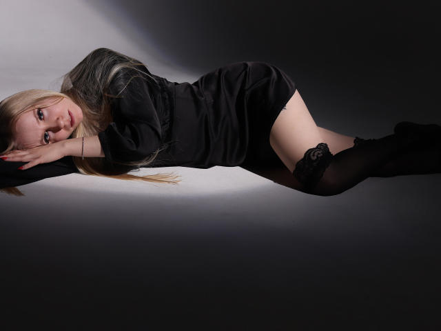 MilanaSun è sdraiata su un fianco, indossa un vestito nero e calze fino alla coscia, con i glutei sollevati e la testa appoggiata al braccio, i capelli biondi le ricadono sulla fronte..