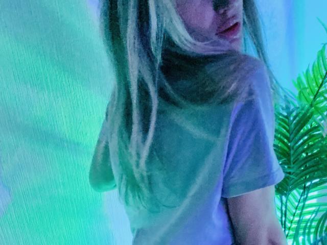 AlicePrings with long blonde hair, looking back, wearing a sheer white shirt, showing her tits, in a green-blue lit room, with a green plant on the right.