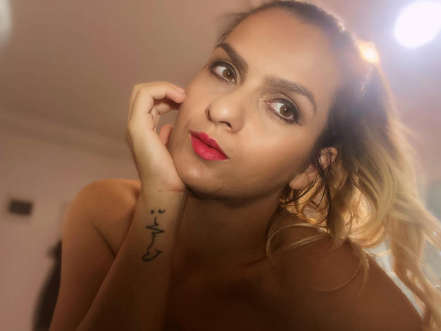 ReveMignoneX has blonde hair in a ponytail, red lipstick, and a tattoo on her left wrist. She's leaning on her hand with a sexy, confident expression.