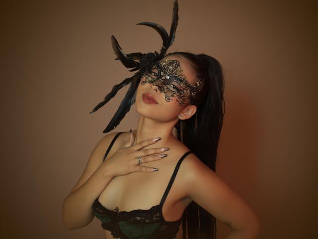 SophieCherryl met een zwart kantmasker, een zwarte veerkroon, een groene kantbeha, een lange zwarte paardenstaart, hand op de borst, geverfde nagels, een ring, en een bruine achtergrond. Borsten, sexy, elegant..