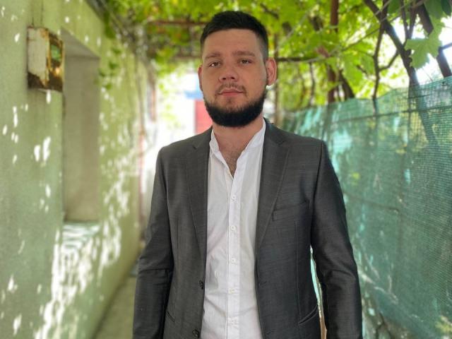 MattShine, un homme barbu avec une courte coupe de cheveux, portant un costume gris sur une chemise blanche, se tient dans un passage couvert, baigné de soleil, verdoyant et ombragé par des feuilles..