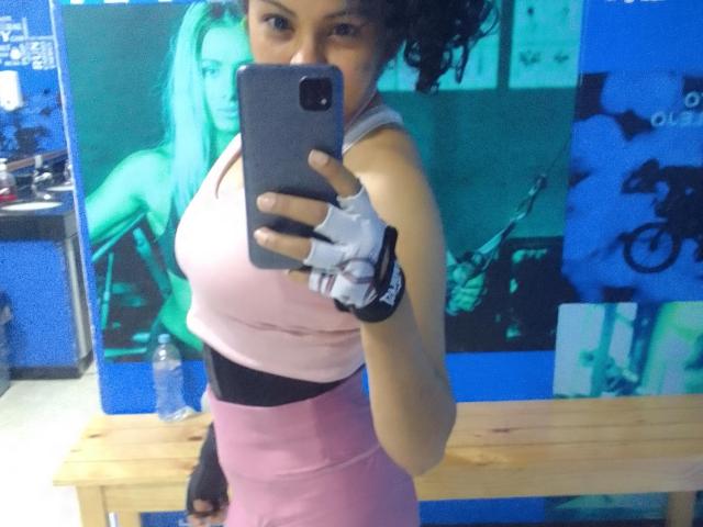 AnnaMesht with curly hair, pink sports top, pink high-waist shorts, white finger glove, holding phone, mirror selfie, visible ass, blue wall, wooden bench, water bottle.