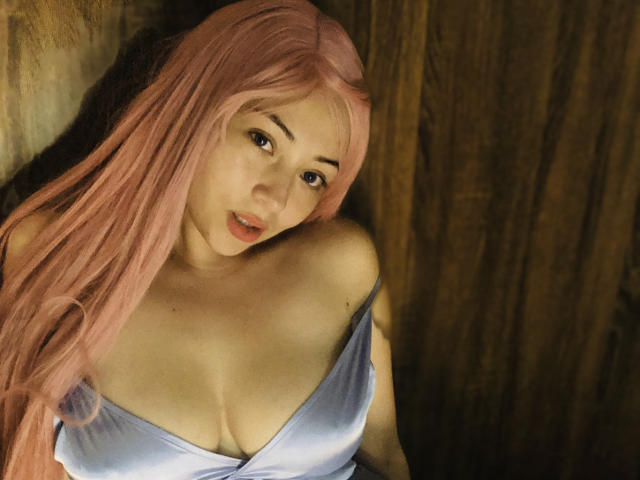 Harumi Noue, com cabelo rosa, olhando para cima com os lábios entreabertos, exibindo seus seios em um vestido azul claro brilhante e com decote baixo. Seus olhos estão ligeiramente abertos e sua expressão é relaxada..