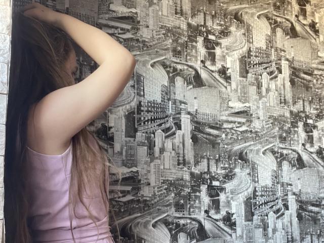 Dooris with long brown hair, wearing a pink tank top, has her hand in her hair, standing in front of a black-and-white cityscape wallpaper, showing urban roads and buildings.