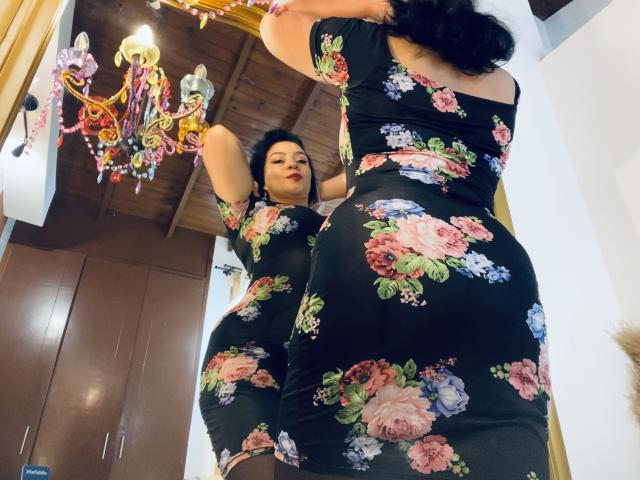 AlisGourmande in a black floral dress, showing off her big ass and small tits in a mirror, with dark hair and red lipstick, standing in a room.