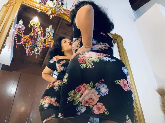 AlisGourmande stands in a floral dress, showing her big ass and large tits, reflected in a mirror, with curly black hair, looking down, in a brightly lit room.