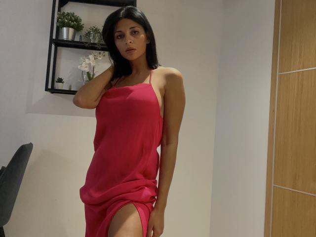 Rodalinda in a pink dress with a thigh slit, black hair, looking serious, hand on shoulder, slim, small tits, white walls, wooden door, shelves with plants in background.