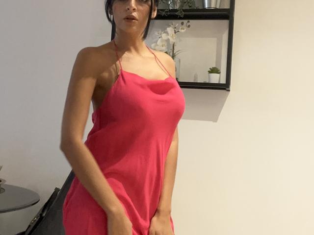 Rodalinda in een roze jurk, die haar borsten en billen laat zien, staat met haar handen in de buurt van haar kruis, draagt een bril, in een kamer met een plank en planten..