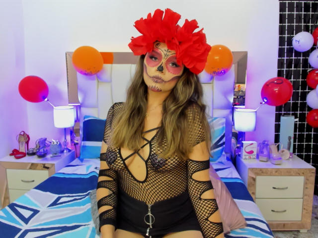 ValerieStone with long brown hair, wearing a red flower mask, black fishnet dress, and black shorts, sitting on a bed with balloons and white lamps in the background.