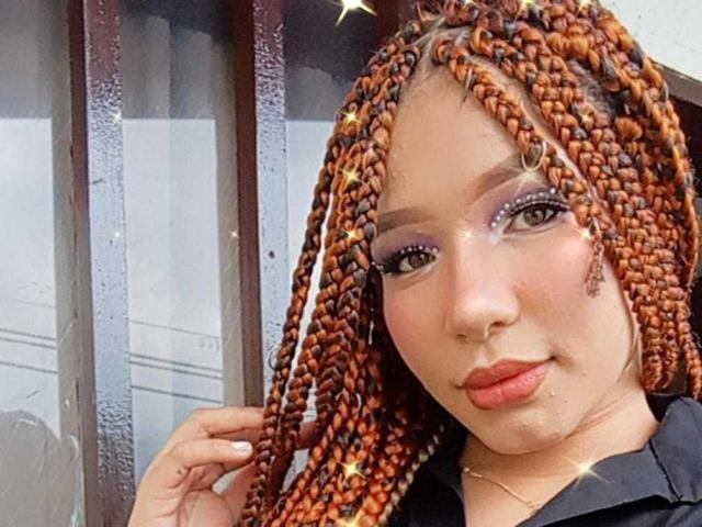 MiaCollins69 tem tranças laranjas, maquiagem roxa nos olhos e lábios vermelhos. Ela está olhando para a câmera com um leve sorriso, segurando o cabelo. Seus seios não estão visíveis..