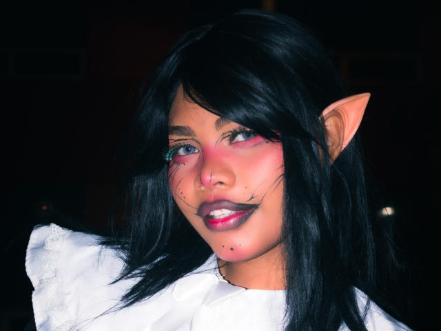 AnniieeBell, con cabello negro, orejas puntiagudas, maquillaje rosa en la cara y una camisa blanca, mostrando una sonrisa linda y ligeramente traviesa, con una pequeña peca negra en la barbilla..