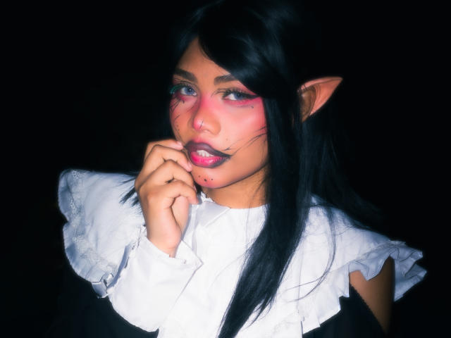 AnniieeBell, con cabello oscuro, orejas puntiagudas, maquillaje rosa en la cara, lápiz labial negro, camisa blanca con volantes y la mano cerca de la boca, luciendo sexy y misteriosa..