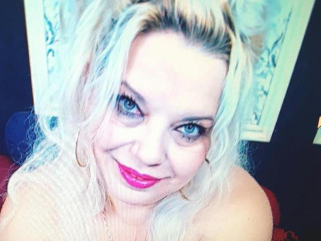 SoniaHotMilf with blonde hair, blue eyes, pink lipstick, and hoop earrings, smiling at the camera, showing her bare shoulders. Her tits are partially visible.