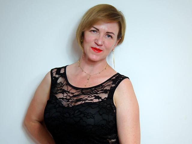 FeliziaStar with short blonde hair, wearing a black lace top, red lipstick, and gold necklace, standing against a white wall, showing her tits and a cute smile.