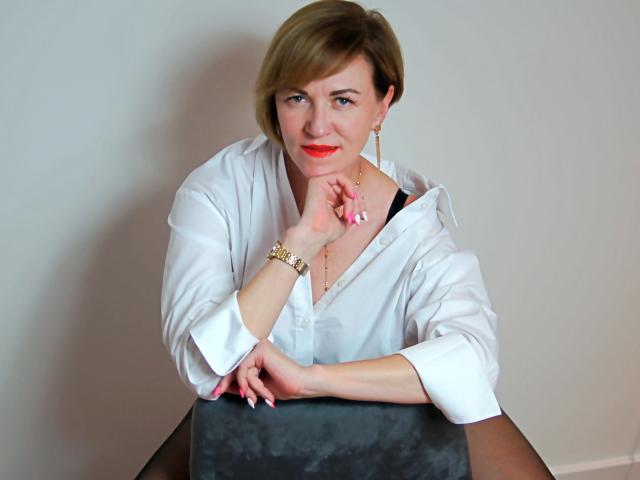 FeliziaStar with short blonde hair, white blouse, red lipstick, gold earrings, and pink nails, leaning on a dark table, smiling slightly, right hand touching chin.