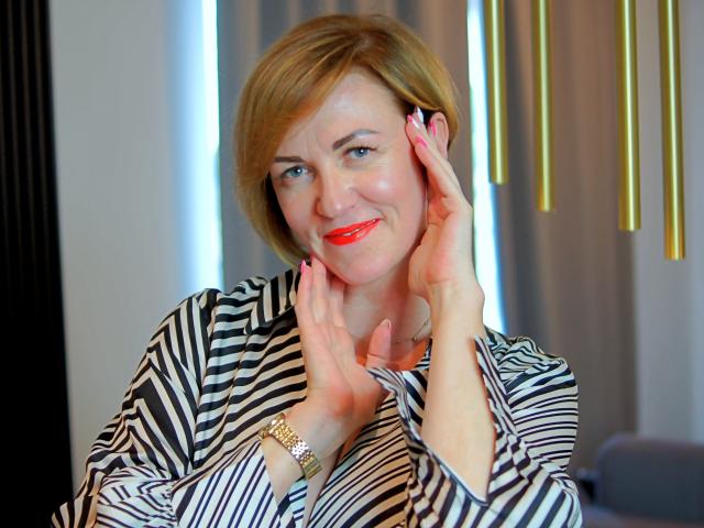 FelisiaStar with short blonde hair, striped shirt, red lipstick, and pink nails, smiling, hands near face, gold watch, and relaxed expression.