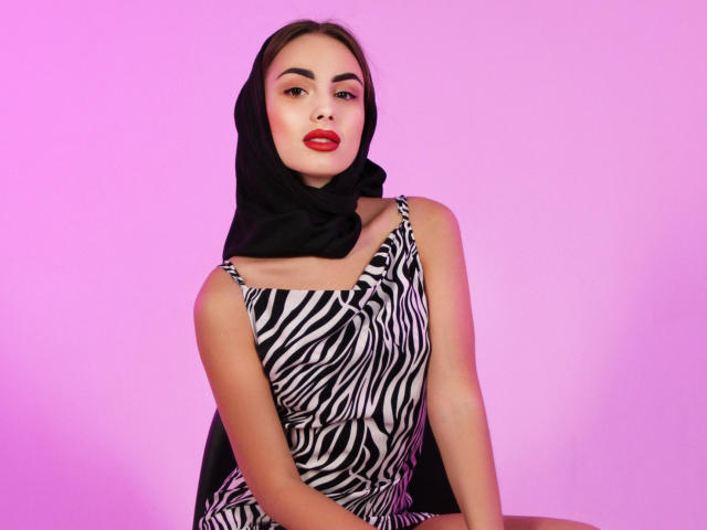 Melisa Wiesel, com lábios vermelhos, hijab preto e vestido listrado como zebra, sentada, mostrando os seios, fundo rosa, sexy, confiante..