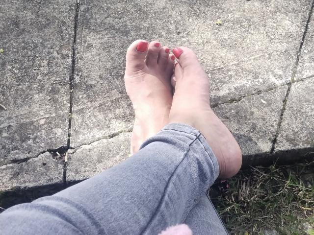 De blote voeten van CoffeCream, met roze geschilderde teennagels, gekruist bij de enkels, draagt grijze jeans en rust op een gebarsten stenen oppervlak met wat groen gras aan de onderkant..
