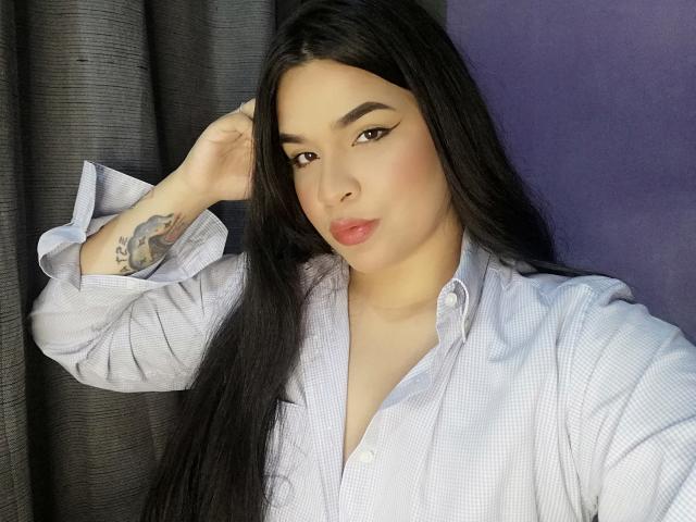 Antonela, con cabello largo y negro, vestida con una camisa blanca a cuadros, tiene un tatuaje en su brazo derecho, ojos marrones y labios rosados, posando con la mano en el cabello..