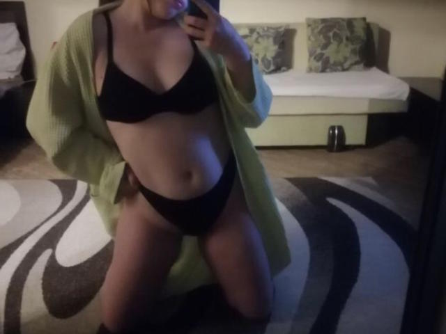 NikiNordic se arrodilla en una habitación, vistiendo un cárdigan verde abierto que revela un sujetador y ropa interior negros, mostrando sus pechos y vulva, con un teléfono en la mano..