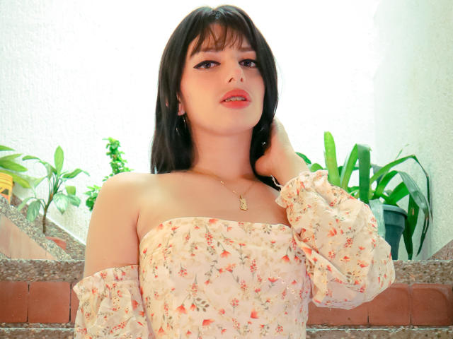 LeticiaFlorenz mit heller Haut, schwarzen Haaren und rotem Lippenstift, trägt ein blumiges Kleid mit einem Schulter frei, eine Hand im Haar, eine Halskette, im Hintergrund grüne Pflanzen, eine Mauer aus Granit und Ziegel..