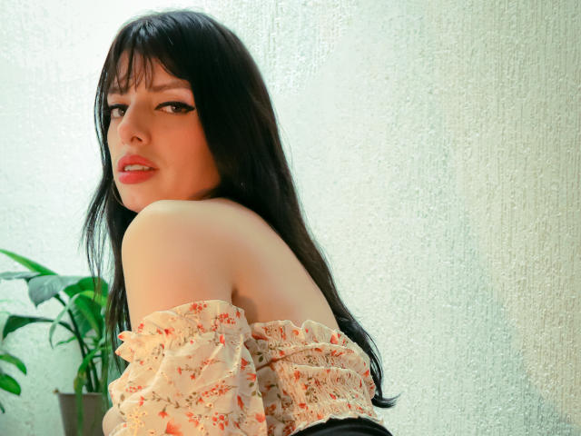 LeticiaFlorenz with long black hair, wearing an off-shoulder floral top, looking over her shoulder, pale skin, red lipstick, green plant in the left, textured white wall.