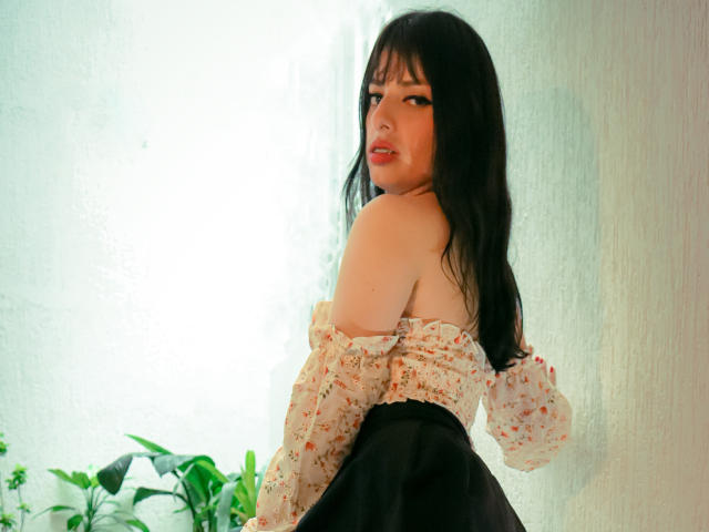 LeticiaFlorenz with long black hair, looking back, wearing an off-shoulder floral top and black skirt, showing her bare shoulders and subtle ass. She's near green plants.