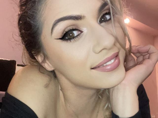 LucilleXSugar a de grands yeux verts avec un trait d'eye-liner noir, un joli sourire avec des lèvres roses et brillantes, et elle repose sa tête sur sa main, portant un haut noir à une épaule..