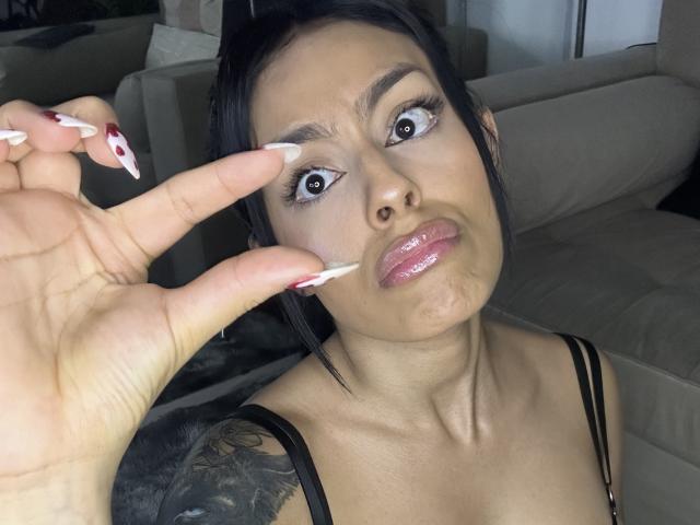 DellaSmith, com uma expressão de choque, cabelo preto, lábios rosados e brilhantes e unhas longas e brancas, tem esperma no rosto, vestindo uma blusa preta com alças, com a mão perto da bochecha..